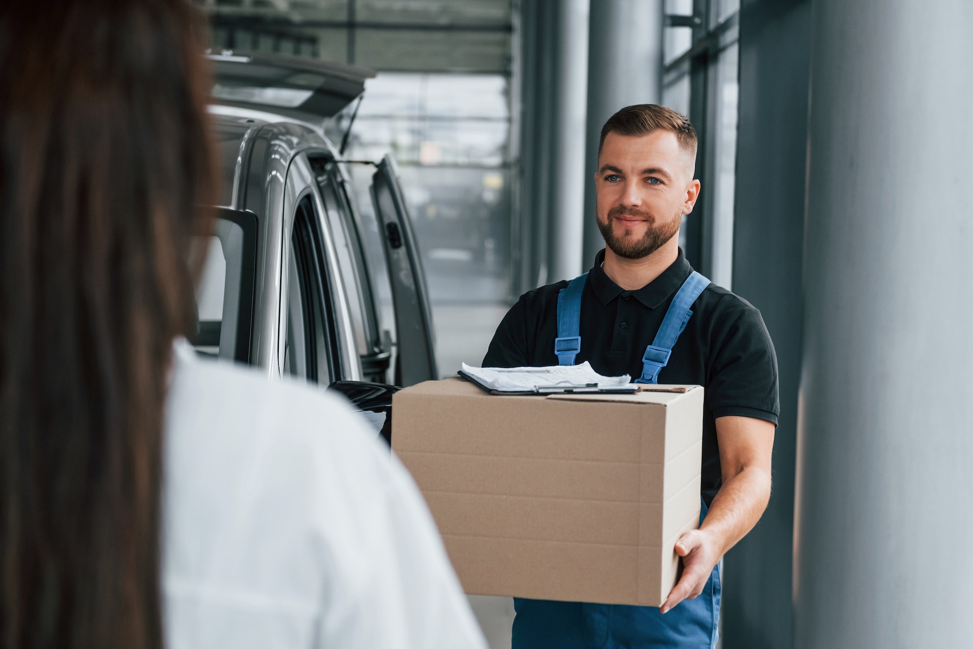 Woman waits for her order. Delivery man in uniform is indoors with car